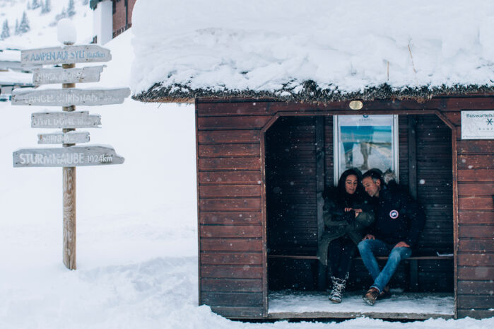 zwei Personen warten in einem holzigen, schneebedeckten Bushäuschen, inmitten der traumhaften Winterlandschaft