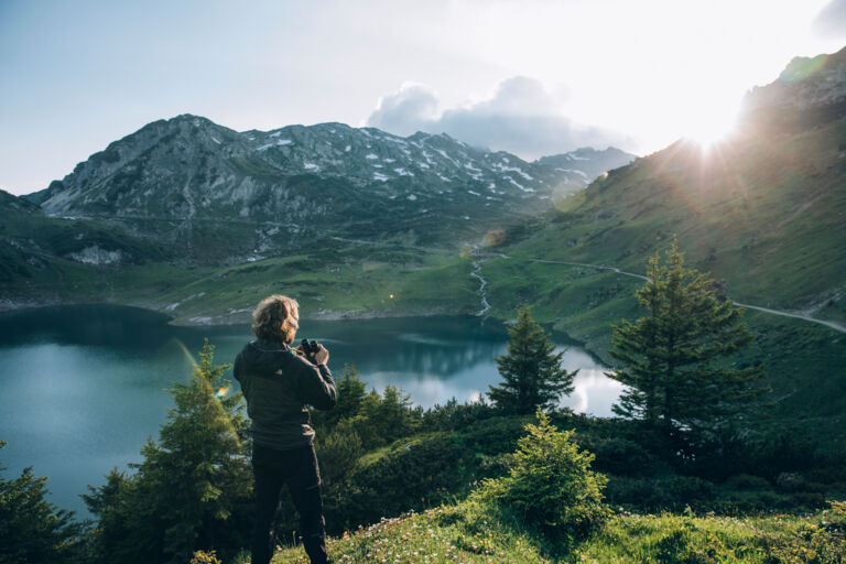 die ersten Sonnenstrahlen scheinen über den Berg zum Formarinsee, welche von einem Wanderer mit der Kamera festgehalten werden
